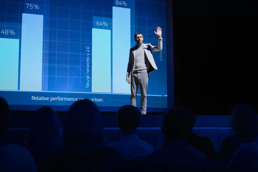 A man presenting on stage with a projection screen displaying relevant content