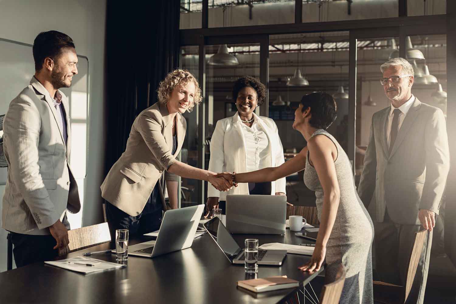 Two businesswomen shaking hands together over a table during a meeting with colleagues in a boardroom in the late afternoon