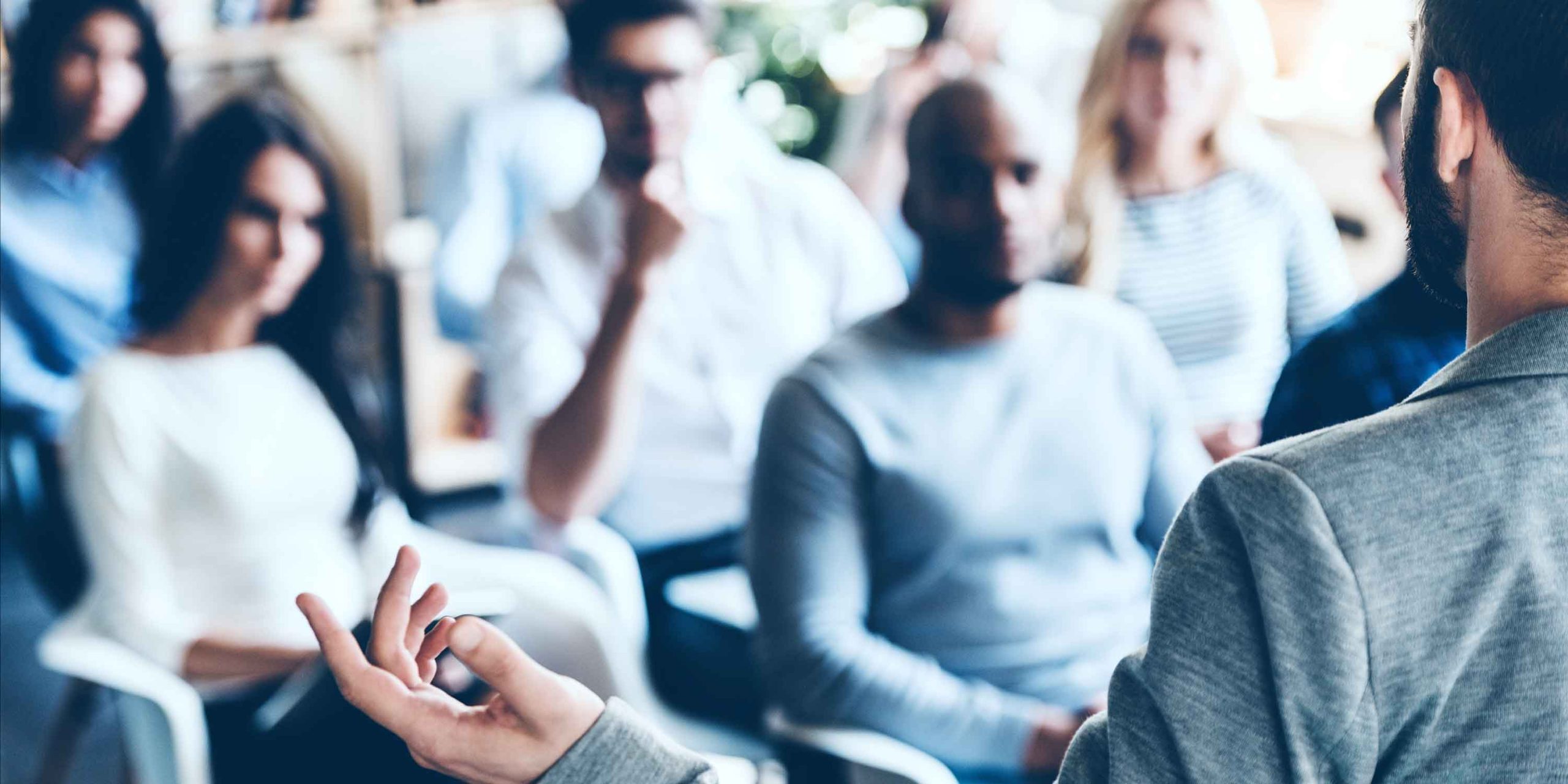 Rear view of man gesturing with hand while standing against defocused group of people sitting in front of him