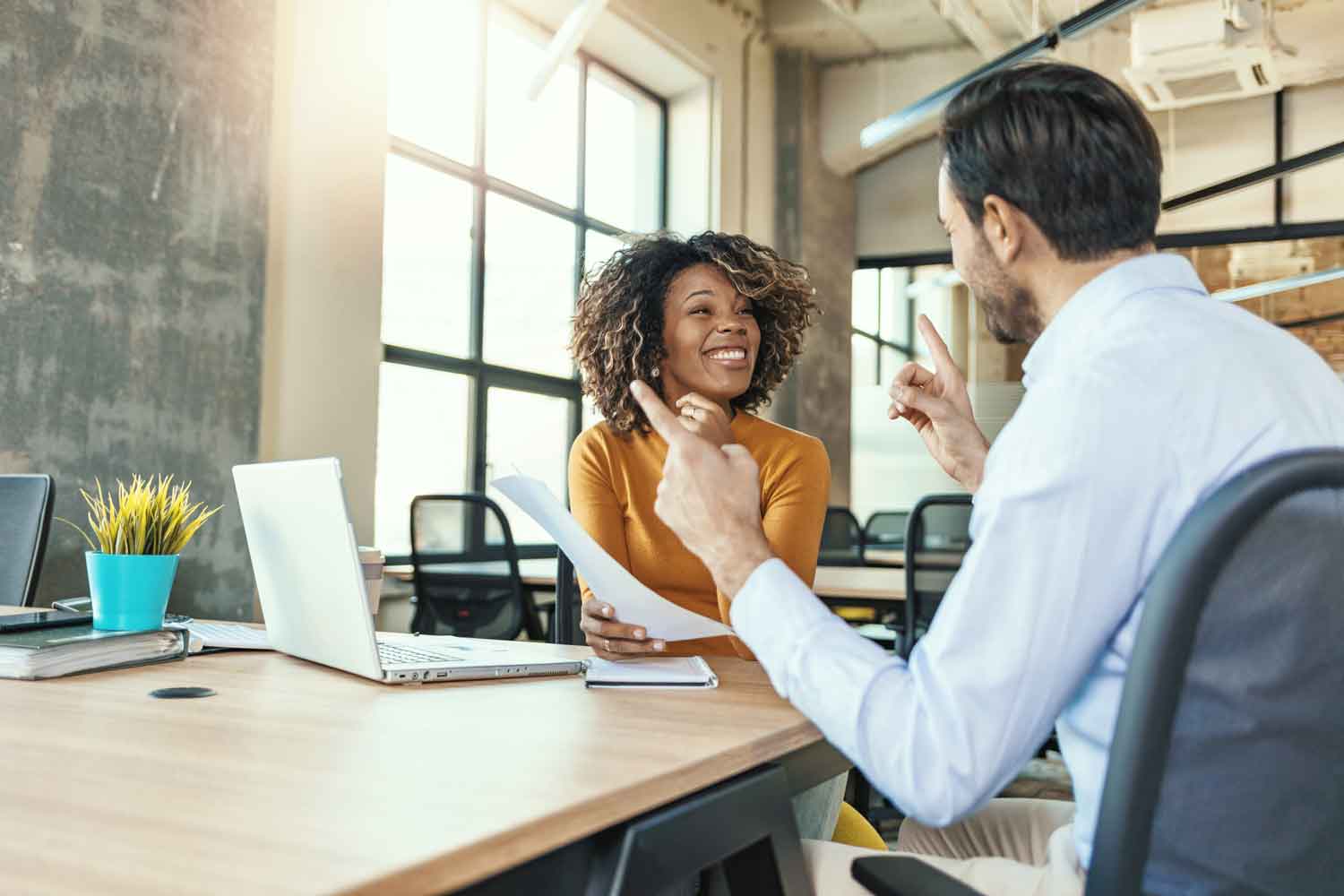 Young woman working with laptop with smile and discussing something with her coworker while sitting at office